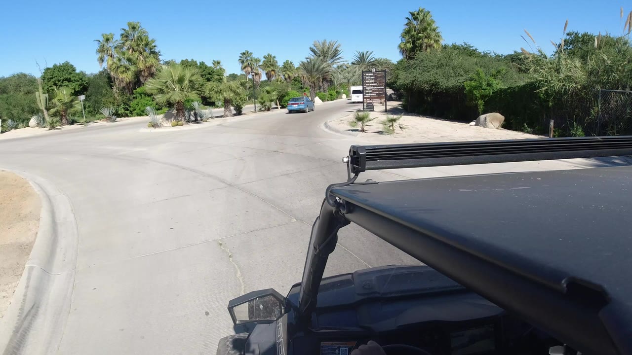 Jeep roof and light bar fill the lower frame as a Los Cabos road curves right past palms, desert shrubs, a blue car, and a dark roadside sign, with no recognizable people visible.