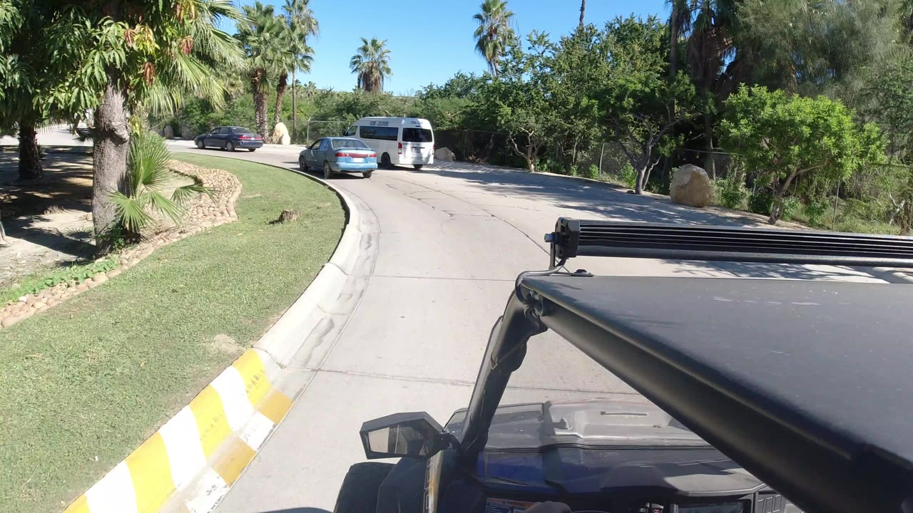 Vehicle-mounted view in Los Cabos with the hood and roof bar in the foreground, a white van and blue car on the road, trimmed grass, palms, and no visible people.