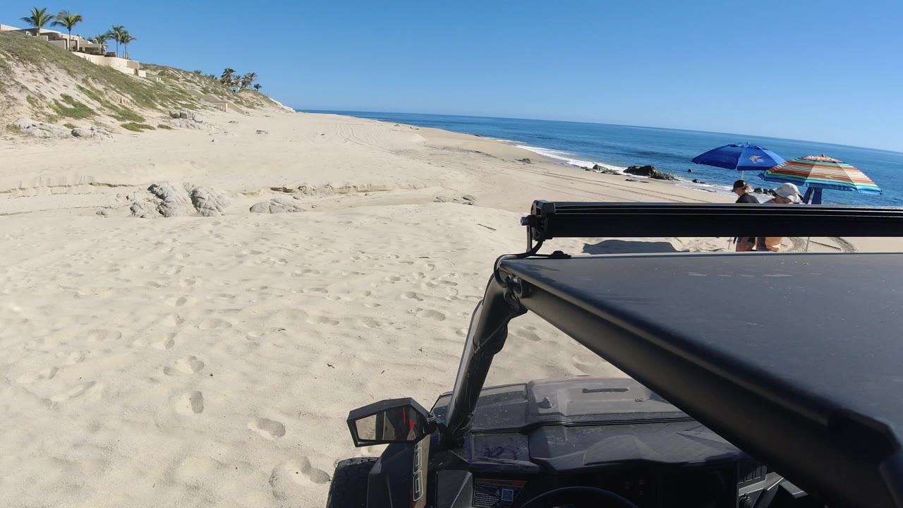 Opening frame in Los Cabos, Mexico, showing a black vehicle in the foreground, pale sand, eroded dunes, blue ocean, and small beachgoers near umbrellas.