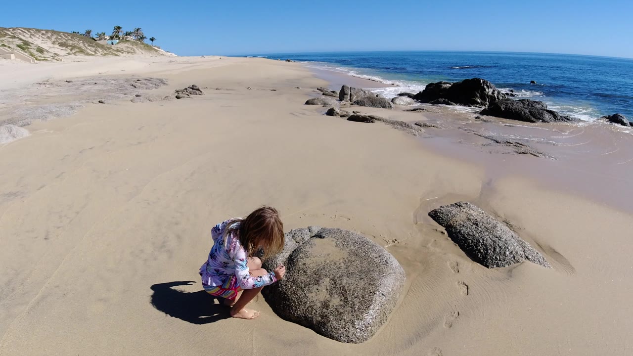 Blonde child crouching beside a rounded rock on a sandy beach.