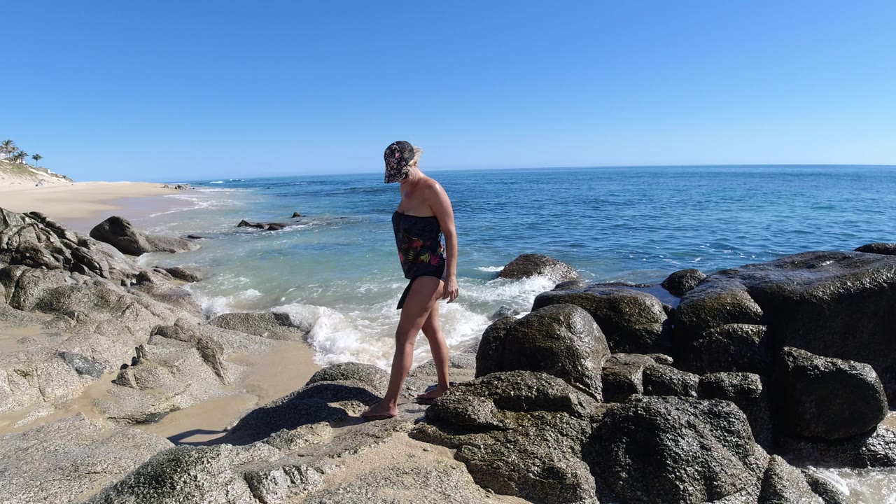 The woman continues across the foreground rocks in Los Cabos, Mexico, while a small wave breaks against stones near the beach.