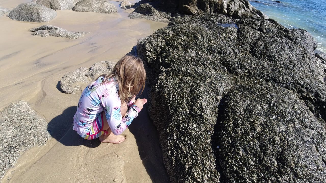 A young girl crouches on pale sand beside large dark rocks on a Los Cabos beach, with bright sun and blue water at the edge of the frame.