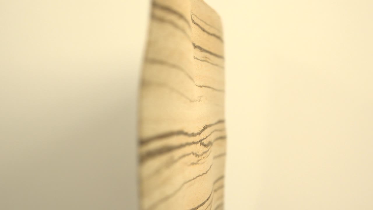 The wooden board edge remains centered between blurred white vertical shapes, emphasizing the dark grain lines and no visible people.