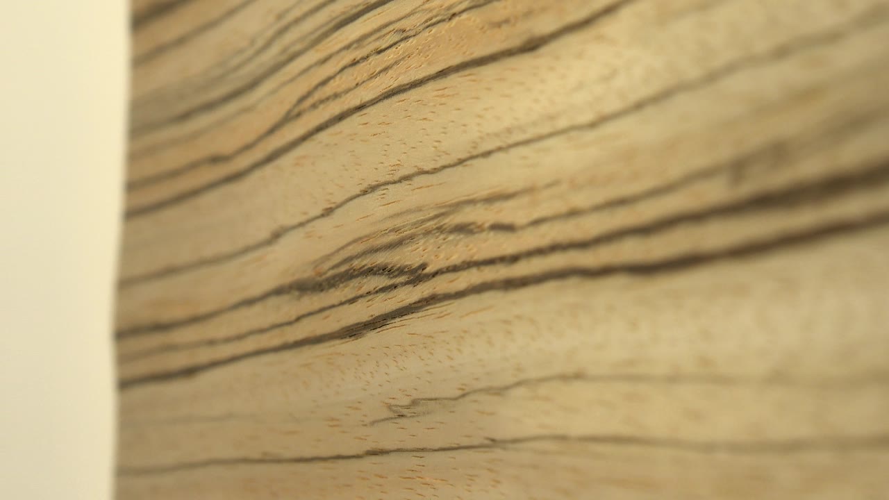 Soft close-up of pale flat wood with dark horizontal grain bands, a blurred off-white vertical edge at left, warm light, and no visible people.