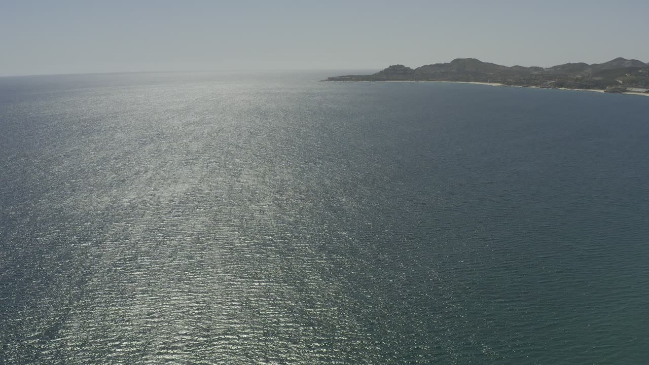 Los Cabos ocean aerial at 0:01, showing textured water, a bright vertical patch of reflected sunlight, distant hills, and no visible people.