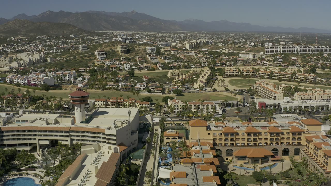 Opening aerial frame over Los Cabos, Mexico, with pale buildings, terracotta roofs, roads, golf fairways, dry hills, and distant mountains under clear daylight, with no visible people.