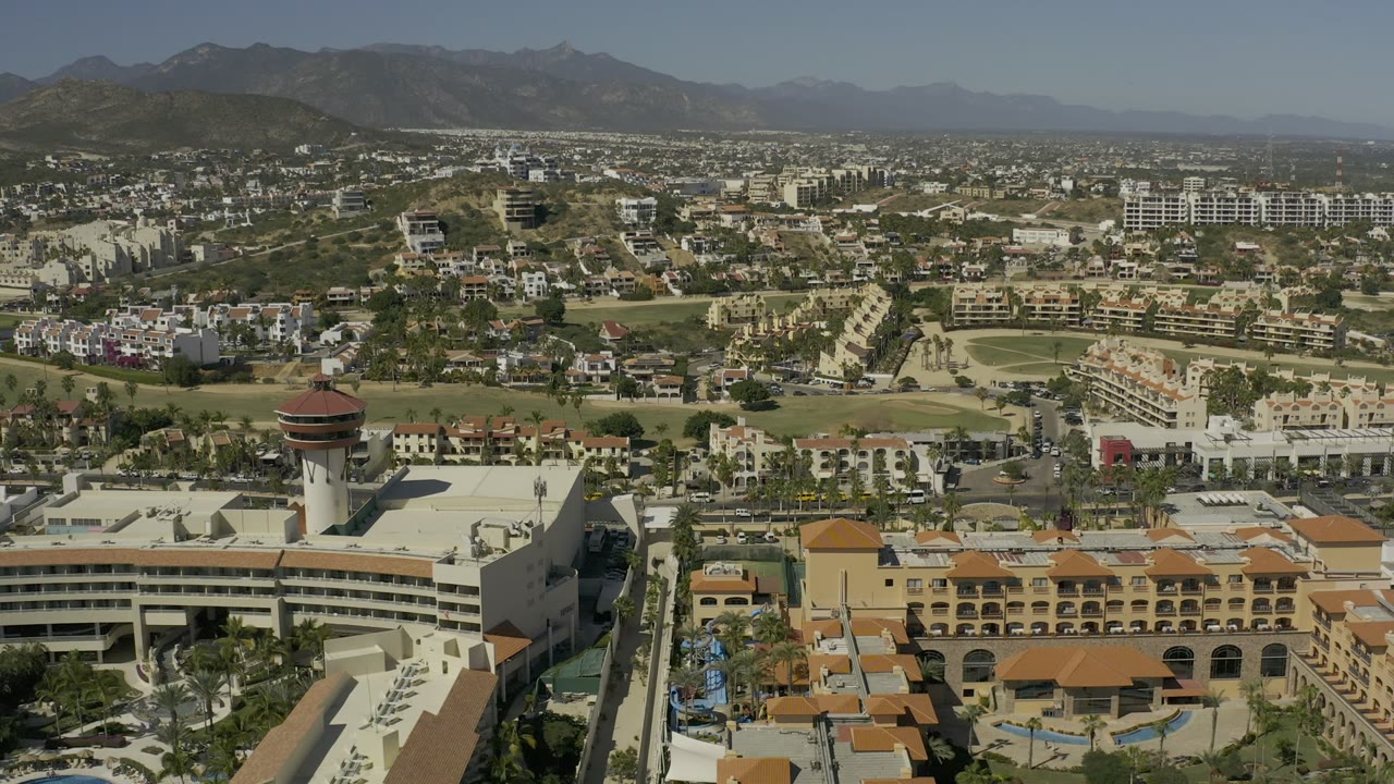 Second aerial frame in Los Cabos showing the round-roofed tower nearer the foreground, resort blocks, road lanes, green fairways, low city development, and mountains, with no visible people.