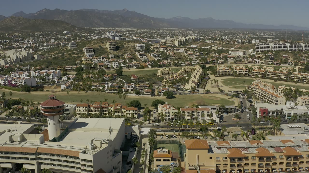 Aerial frame in Los Cabos, Mexico, showing the foreground tower slightly nearer center, cream buildings, road grids, hillside housing, mountain ridges, and no visible people.