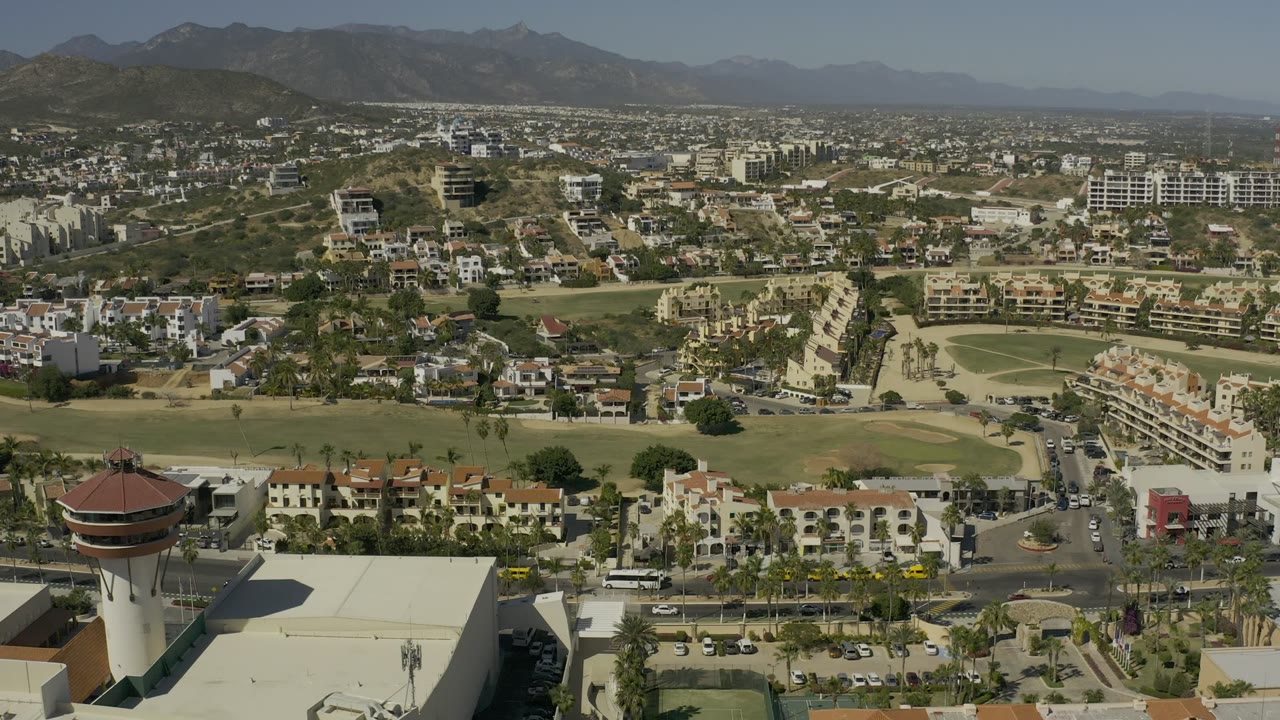 Opening aerial frame over Los Cabos, Mexico with a round tower-like building at lower left, palm-lined roads, green open grounds, hillside homes, mountains in the distance, and no visible people.