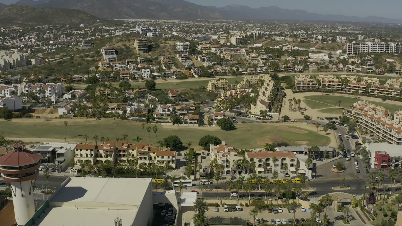 Middle frame over Los Cabos, Mexico with terracotta-roof buildings in the lower frame, curving roads, green grounds, hillside construction, mountain ridges, and no visible people.