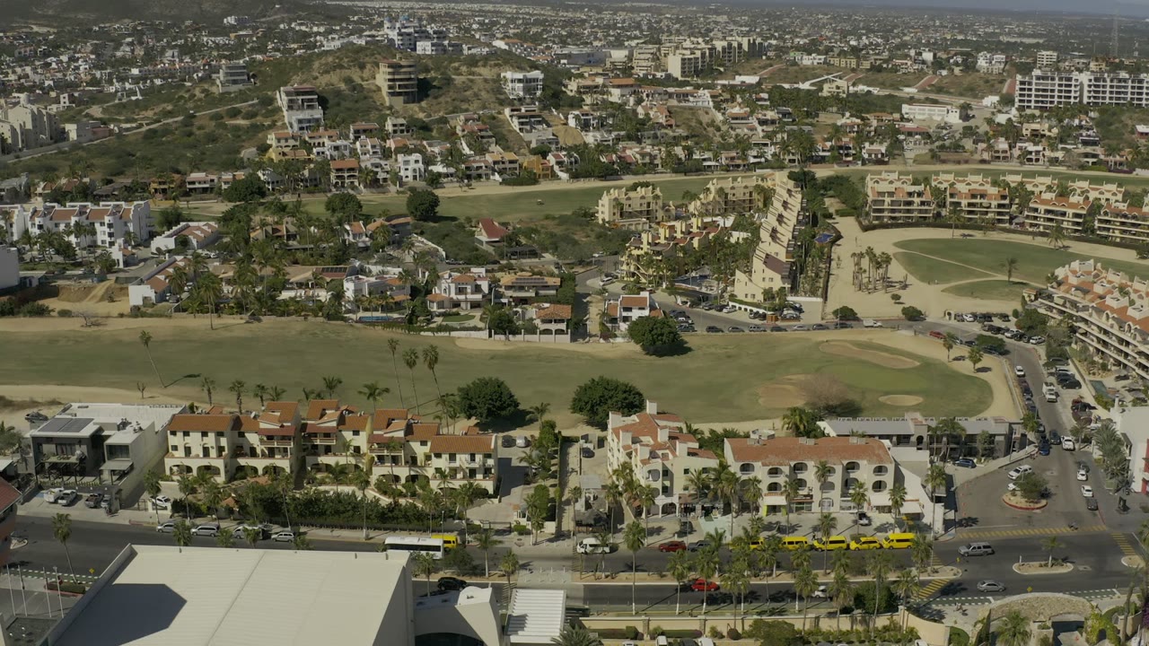 Opening aerial frame over Los Cabos, Mexico, showing a broad foreground road, yellow taxis, red-tile roofs, golf-course greens, hillside homes, and no visible people.