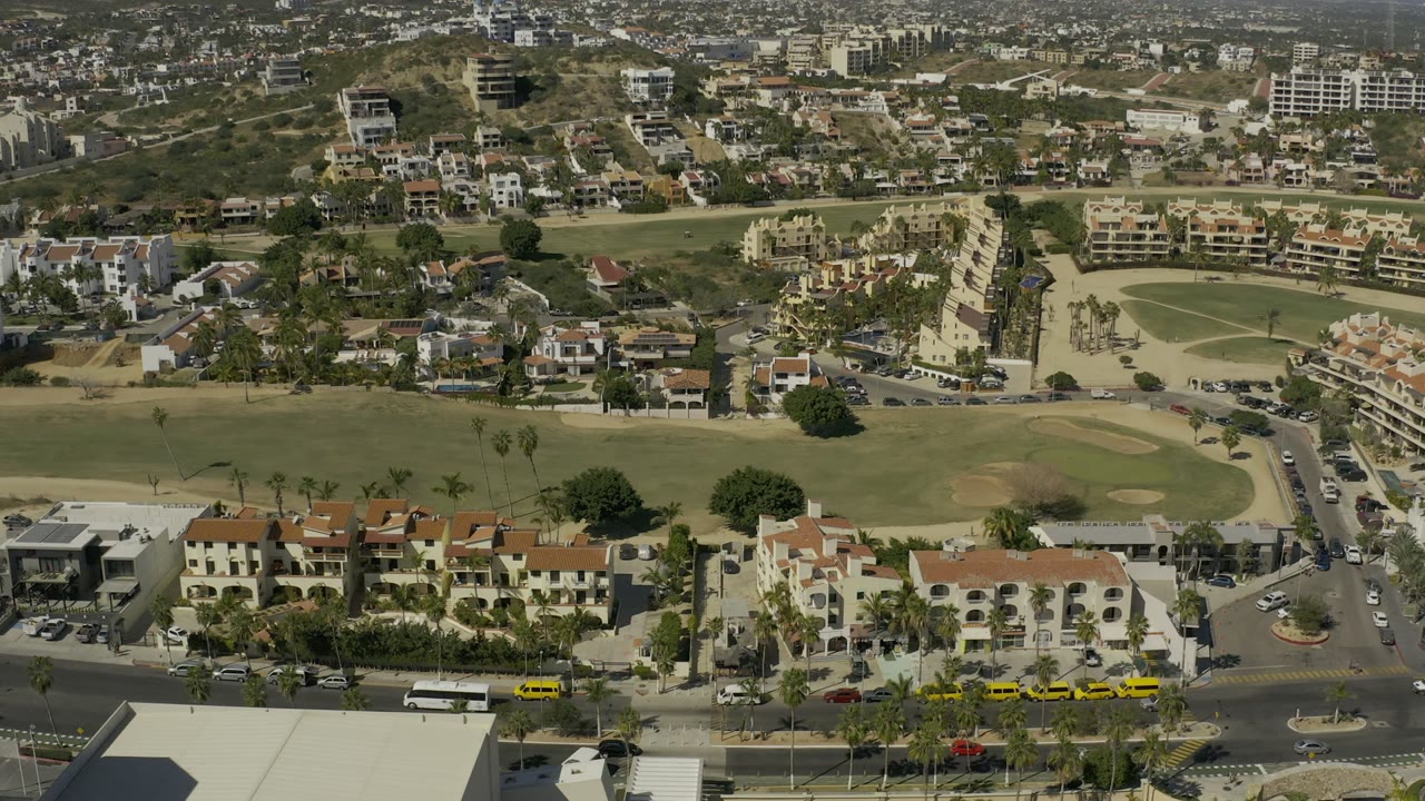 Second aerial frame in Los Cabos, Mexico, with the foreground road crossing below apartment-style buildings, fairways, sand bunkers, palms, parked cars, and no visible people.