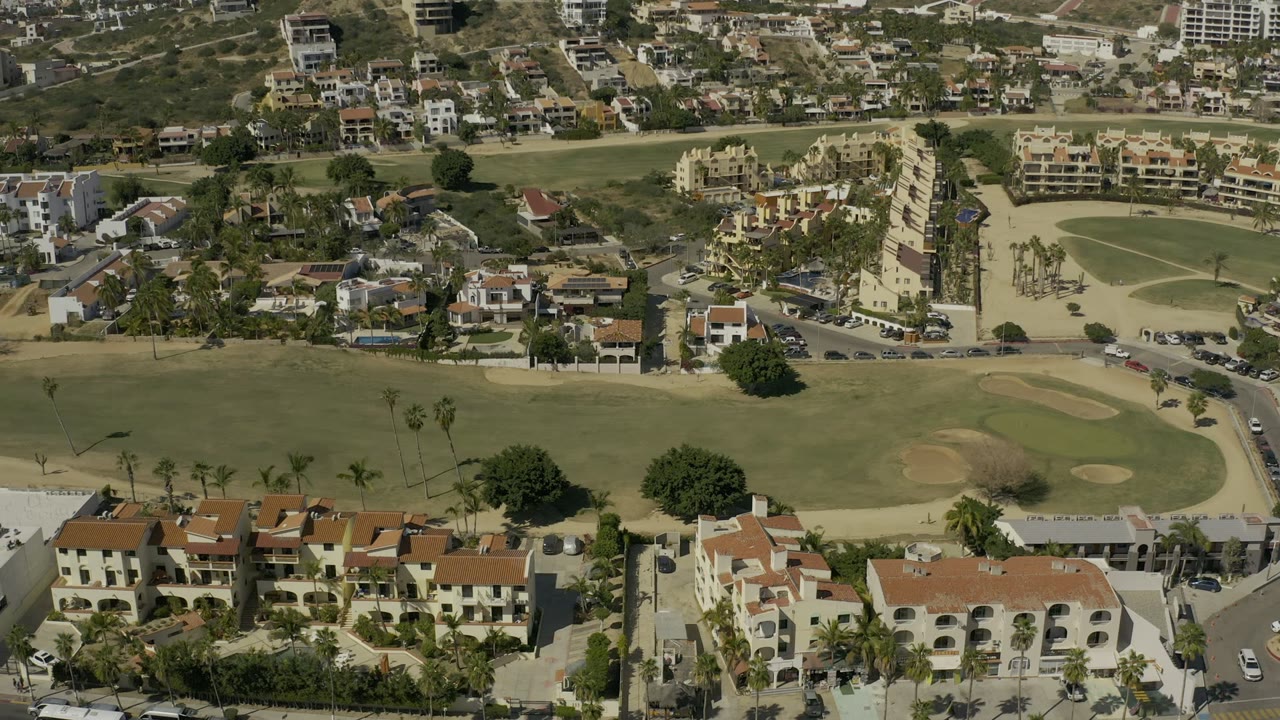 Opening aerial frame over Los Cabos showing a golf fairway with sand bunkers, palm trees, nearby roads, and terracotta-roof residences, with no visible people.