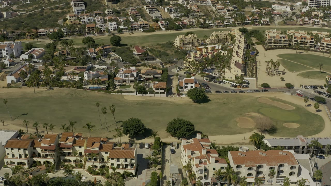 A second aerial frame in Los Cabos holds on golf greens and tan bunkers bordered by cream buildings, palms, roads, and scattered parked cars, with no visible people.