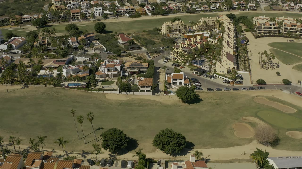 Opening aerial frame over Los Cabos, Mexico, with a golf fairway in the foreground, sand traps, palms, residential streets, and no clearly visible people.