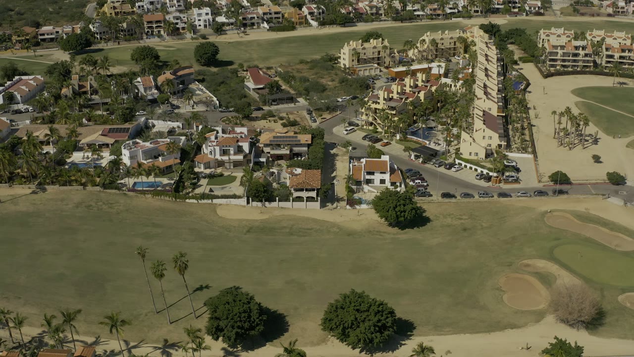 Aerial view in Los Cabos, Mexico, showing more of the fairway and surrounding homes, with apartment buildings and parked cars beyond the course.