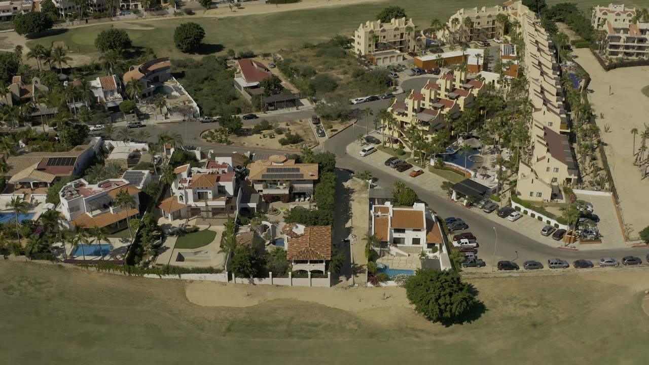 Second aerial frame in Los Cabos, Mexico, with villas and larger cream buildings arranged along paved roads, a fairway at the bottom, pools, palms, and no visible people.