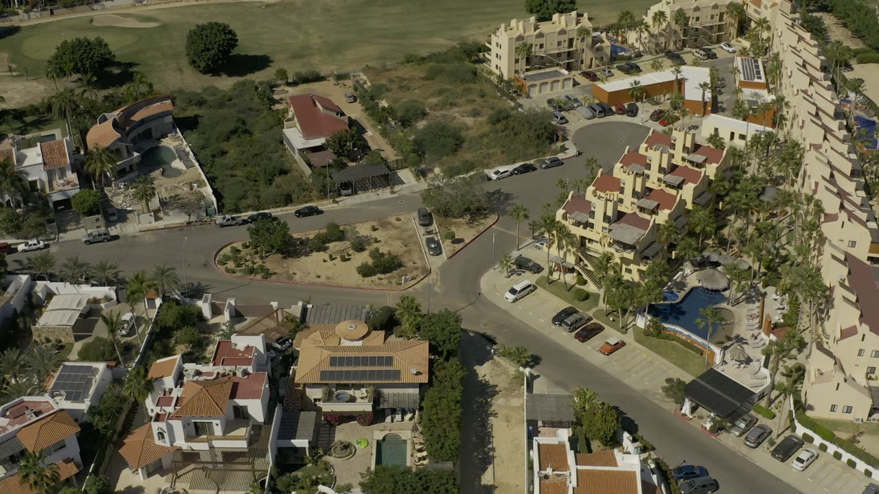 Aerial Los Cabos frame showing a road intersection beside private residences, landscaped palms, blue pools, parked cars, and dry open lots, with no clearly visible people.