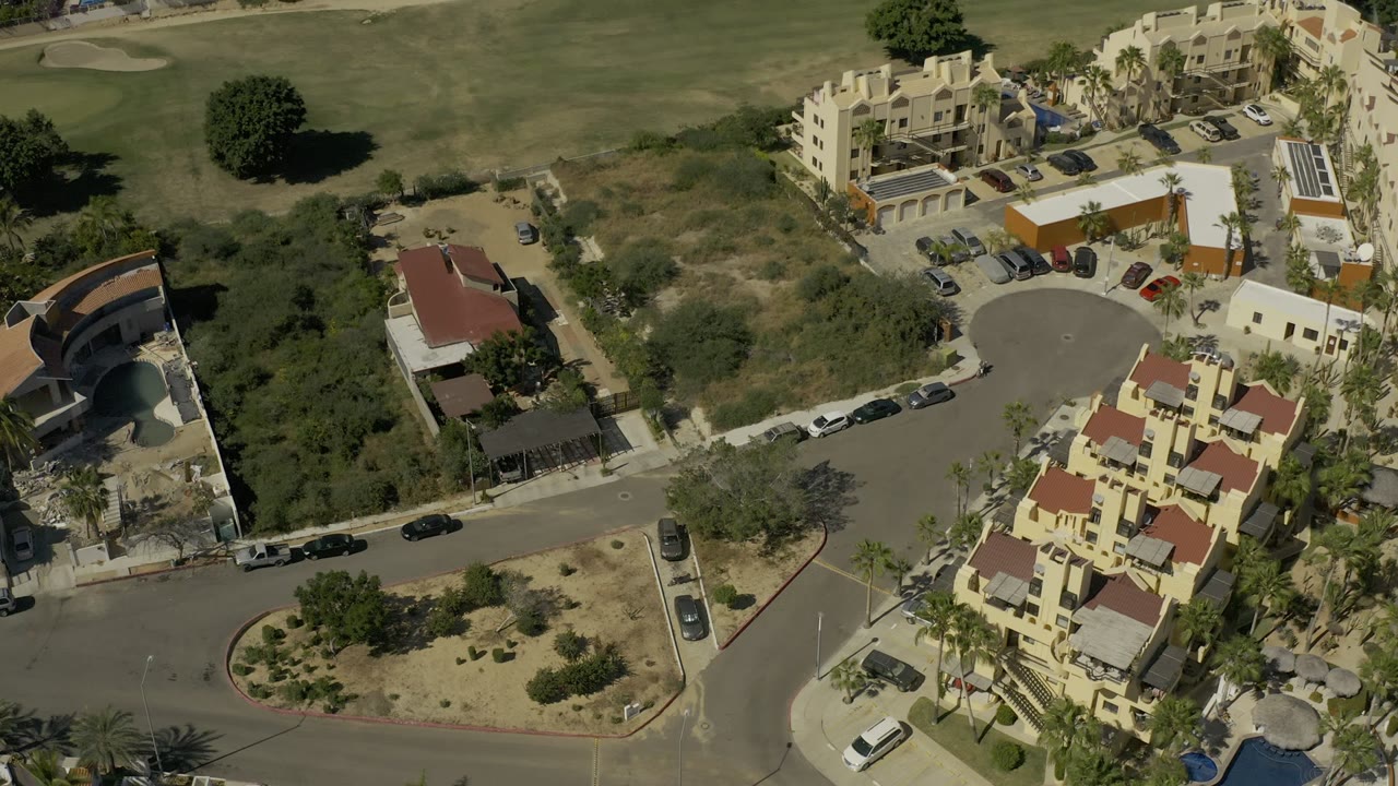 High aerial frame in Los Cabos, Mexico showing cream residential buildings, red roofs, parked cars, palms, dry lots, and green golf course grass with no visible people.