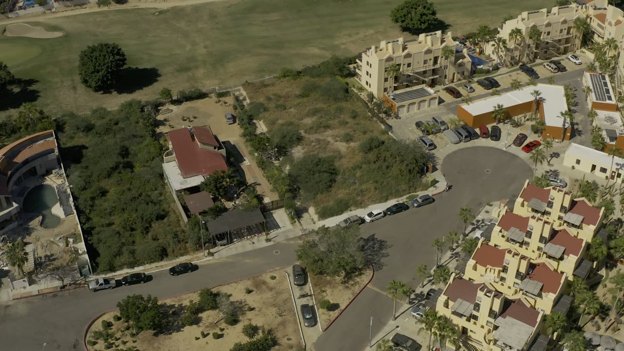 Aerial frame in Los Cabos, Mexico with apartment-style homes, a circular road end, parked vehicles, palms, and golf course turf filling the upper background, with no visible people.