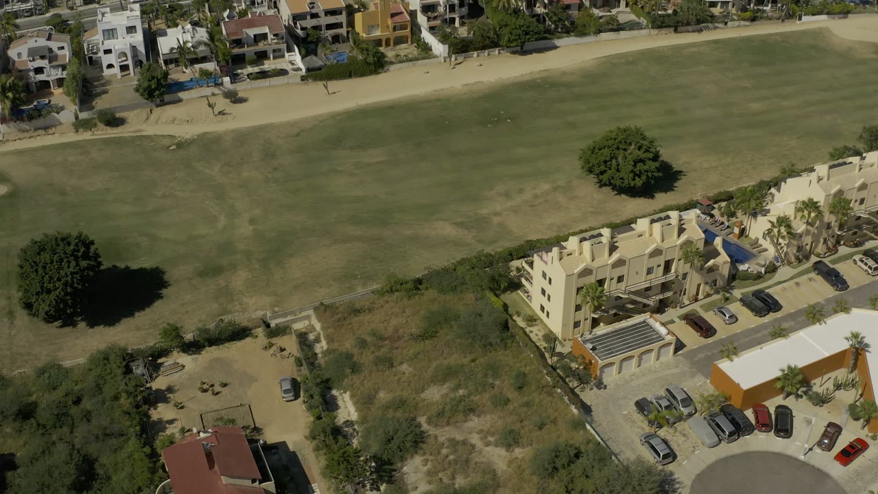 High-angle aerial view in Los Cabos, Mexico, showing a green field below private homes, streets, trees, parked cars, and tiny distant possible people near the path.