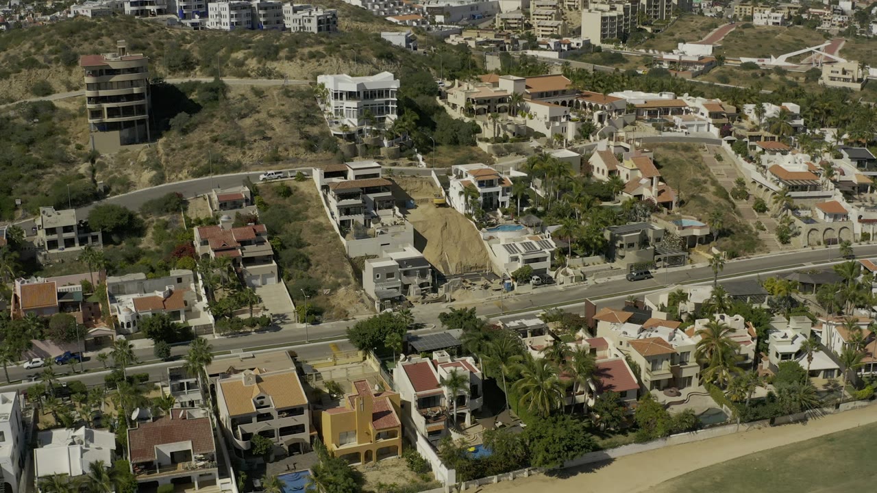 Aerial view in Los Cabos, Mexico with white and terracotta-roofed homes, roadside cars, palms, dry hillside lots, and no visible people.