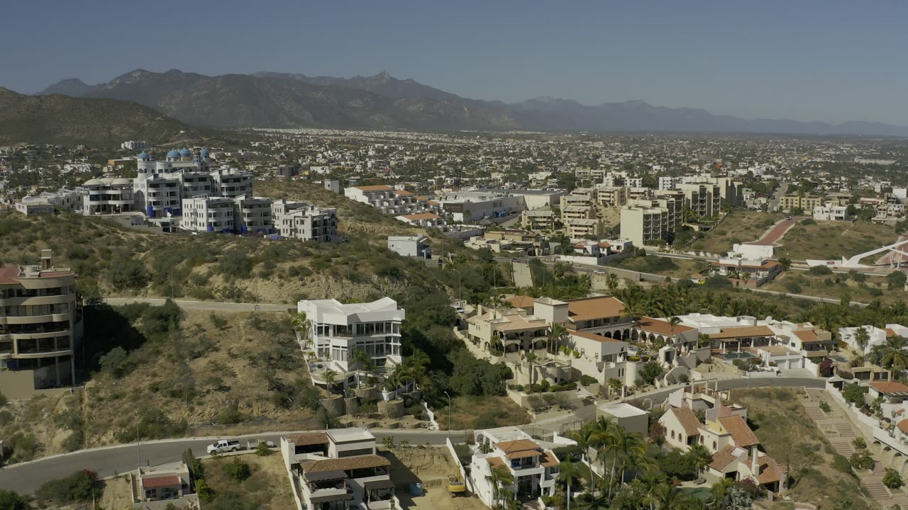 Elevated Los Cabos view with white hillside residences in the foreground, apartment-style buildings across the slope, a spread of city blocks, and no visible people.