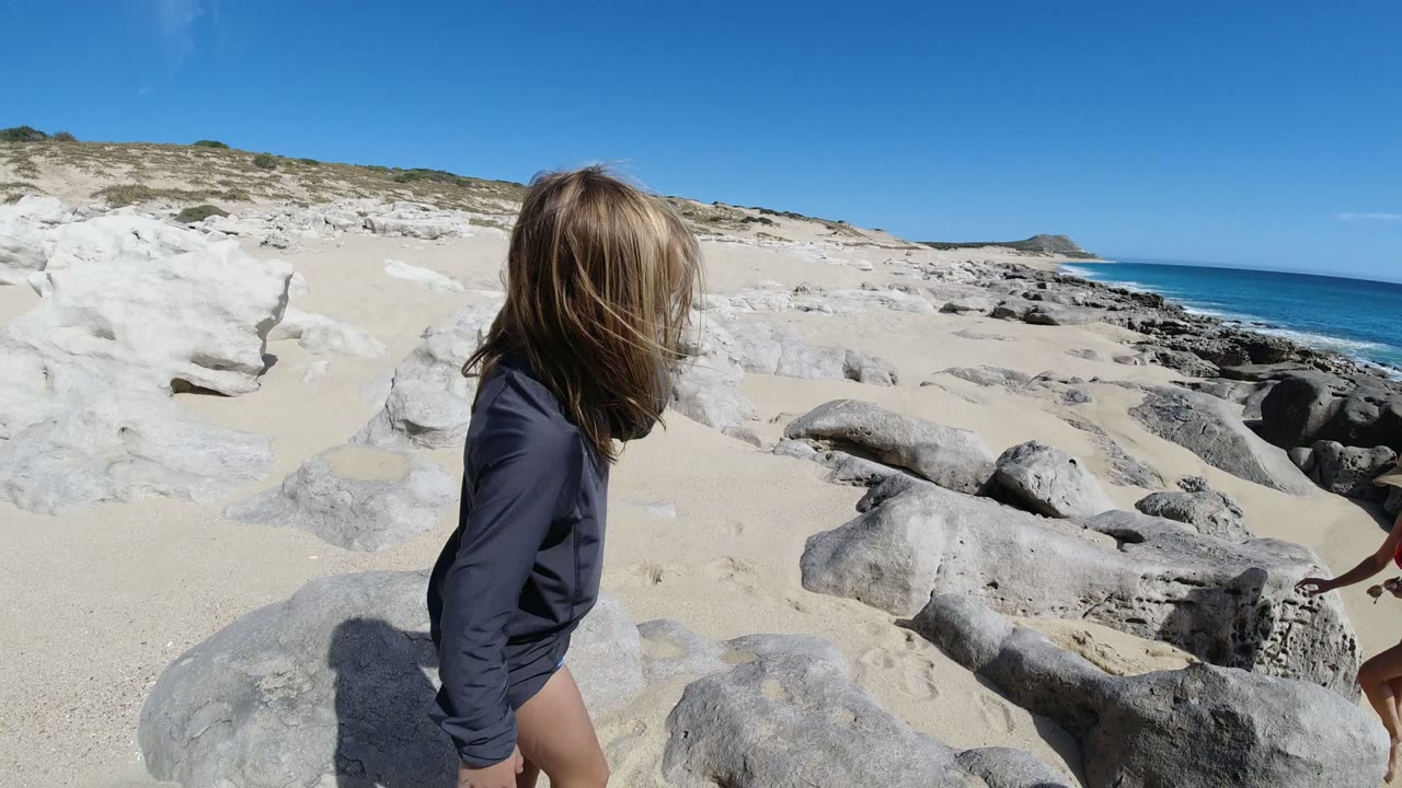 Blonde child standing by pale rocks on a sunny beach.