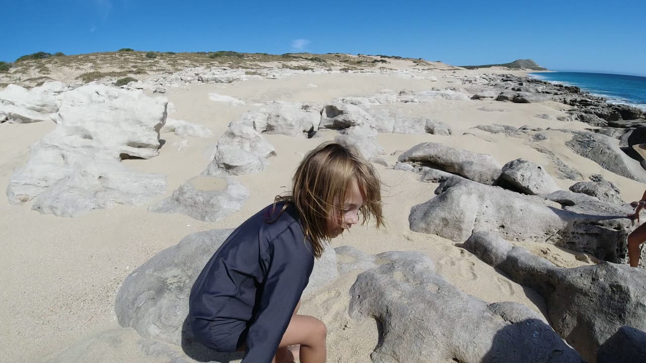 The blonde child crouches lower near large pale rocks on the San Jose Del Cabo beach, with open sand and the blue ocean visible behind the rocky midground.