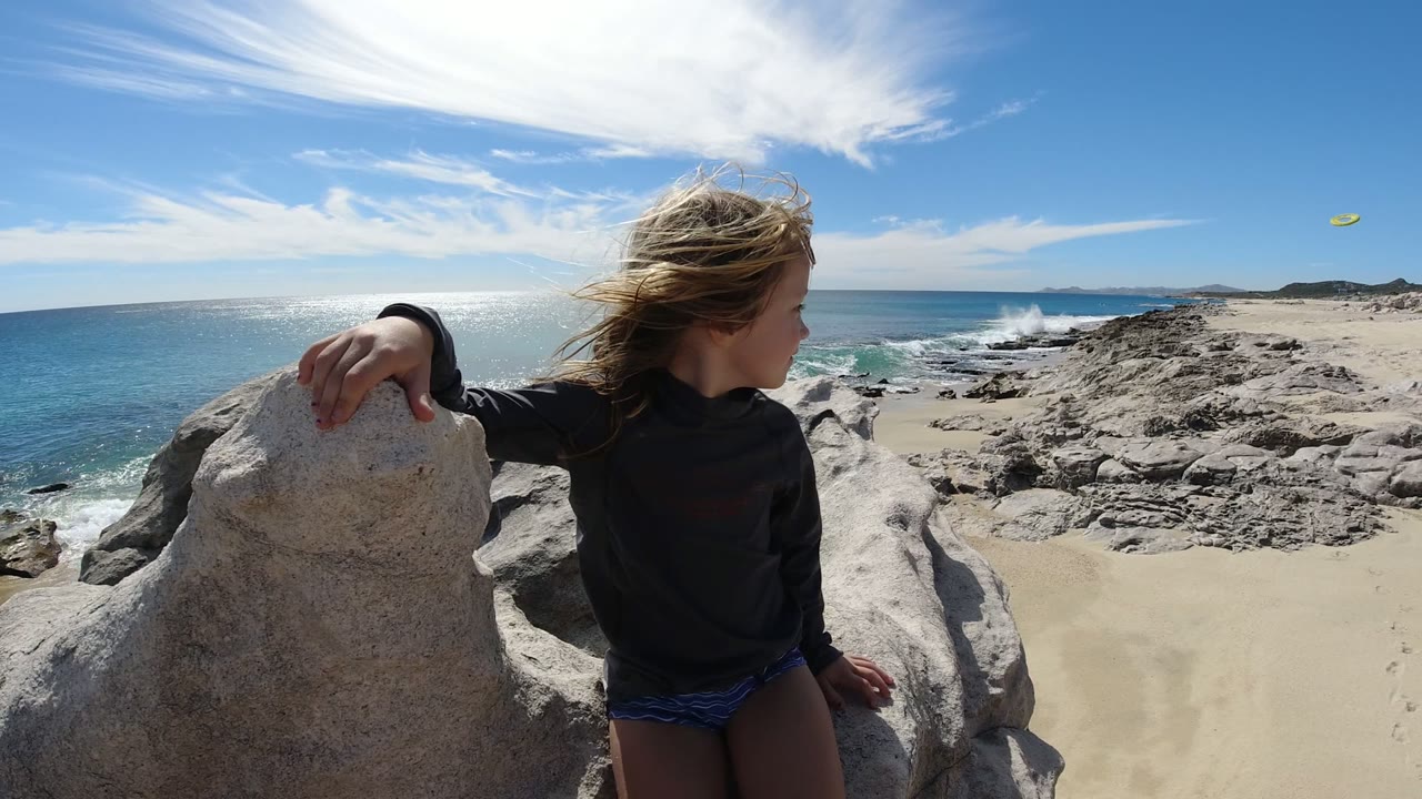 A blonde child in a dark top leans beside a pale rock on a San Jose del Cabo beach, looking toward the shoreline as waves break below.