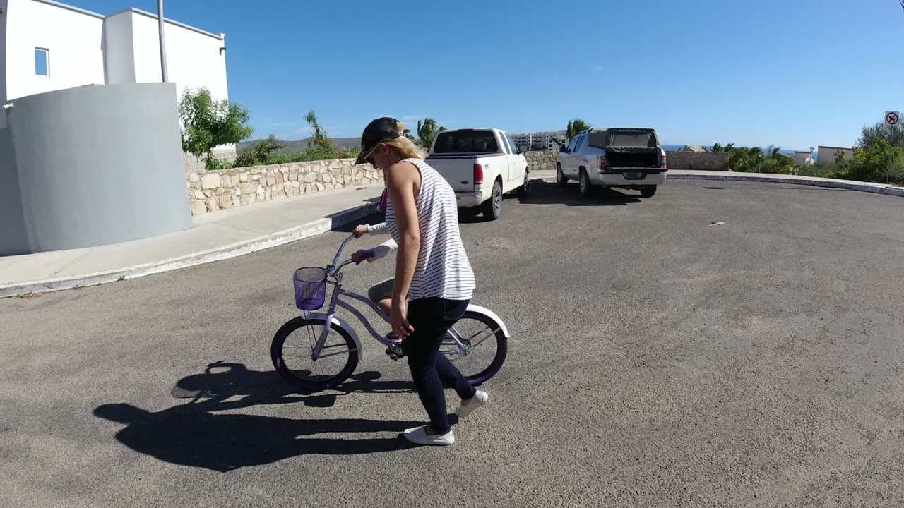 A woman walks next to a young girl on a purple bicycle in San Jose del Cabo, Mexico, with parked pickup trucks, white buildings, and strong shadows on the road.