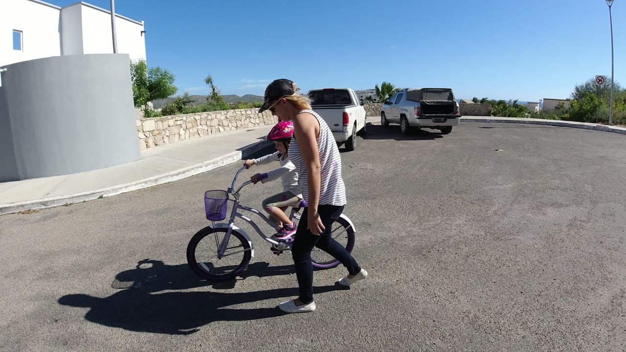 The young girl pedals the purple bicycle while the woman stays close at her side in San Jose del Cabo, Mexico, with the white pickup and stone wall farther back.