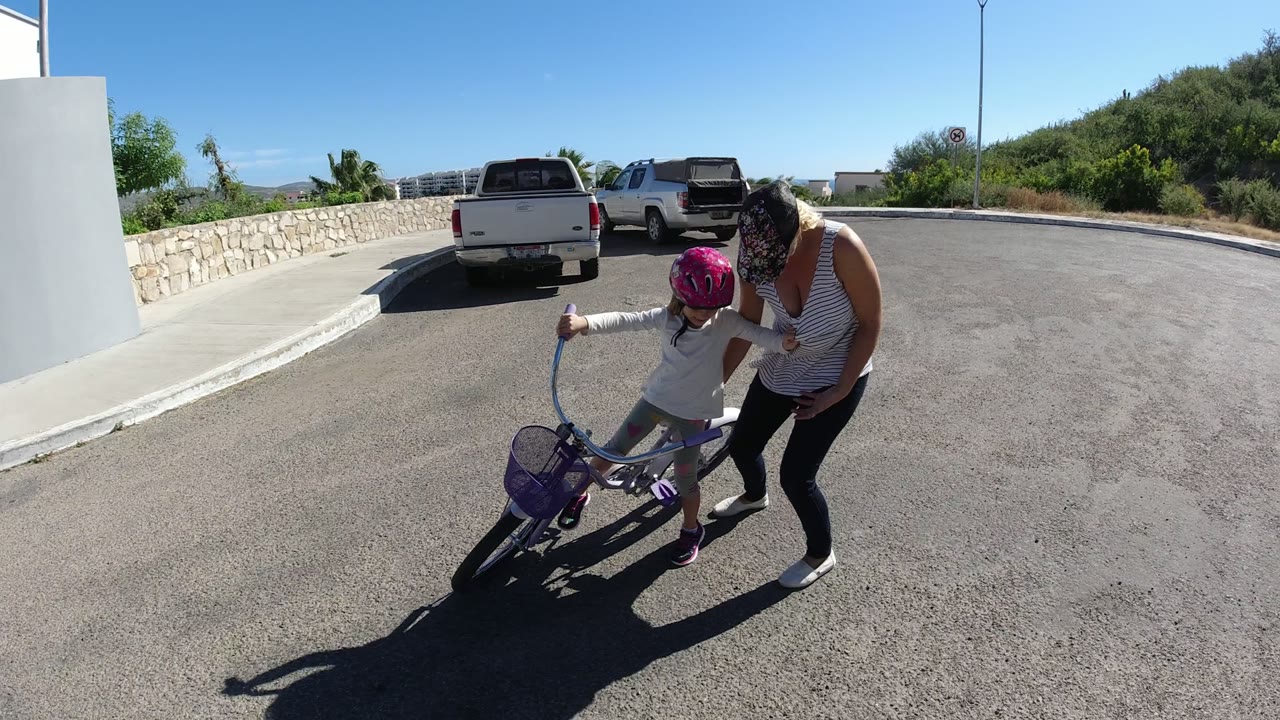 In San Jose del Cabo, a woman bends toward a young girl in a pink helmet as the child handles a small purple bicycle near white walls and parked pickup trucks.