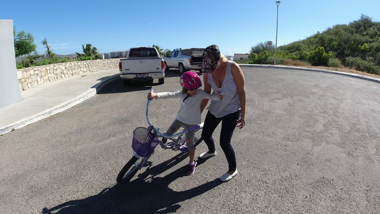 In San Jose del Cabo, the child leans into the handlebars of the purple bicycle while the mother stands to her right on the open paved road.