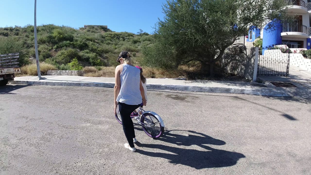 In San Jose del Cabo, the woman and child move farther into the frame with the bicycle angled forward near a curb, scrubby hillside, and blue-and-white residence.