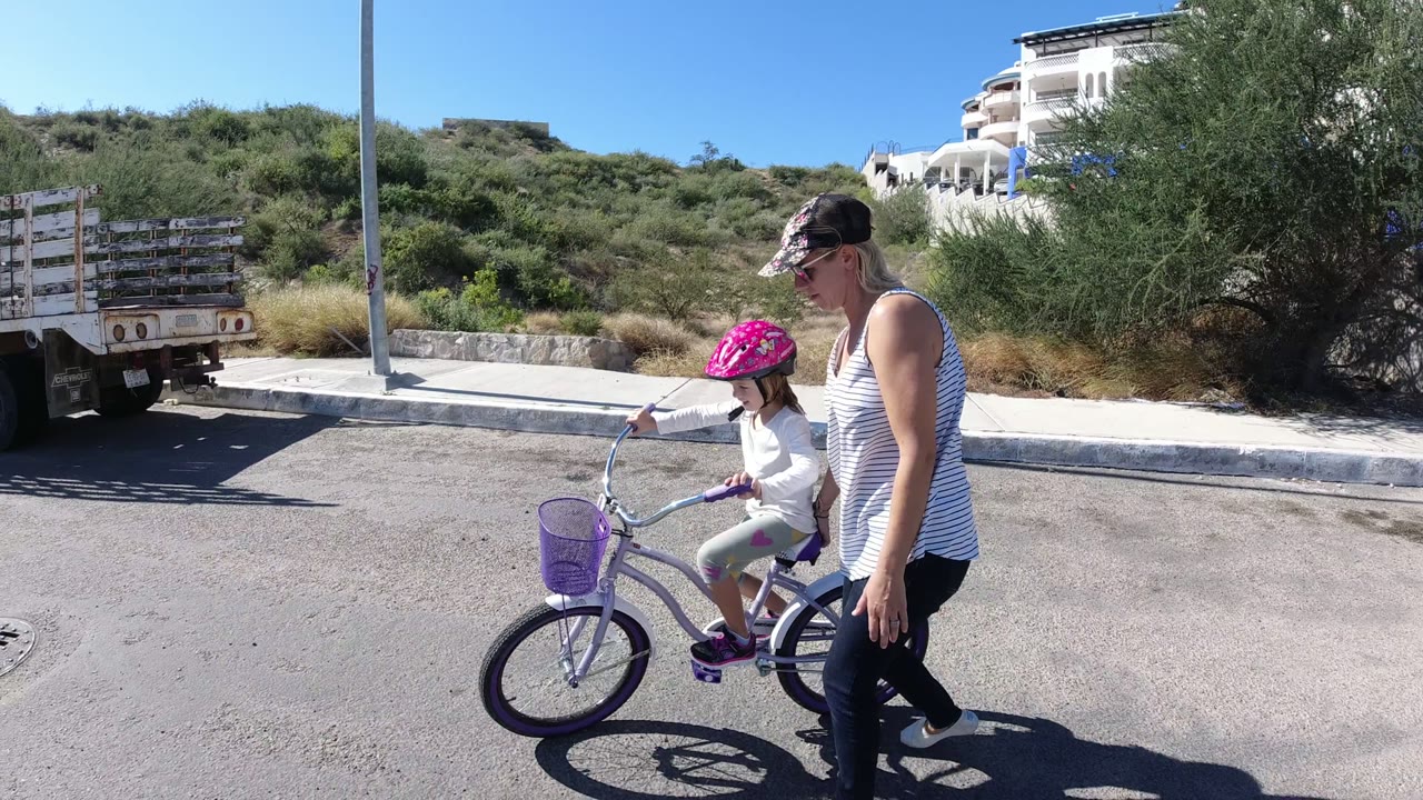 In San Jose del Cabo, Mexico, a woman walks close to a child in a pink helmet on a purple bicycle, with a flatbed truck, hillside brush, and white buildings behind them.
