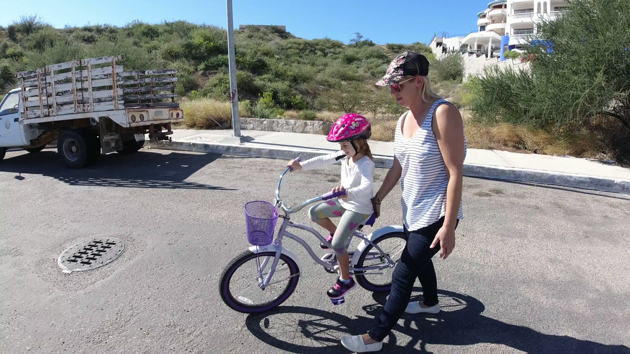 The child rides farther into the frame beside the woman in San Jose del Cabo, Mexico, with the purple basket, bright road surface, curb, hillside, and parked truck visible.