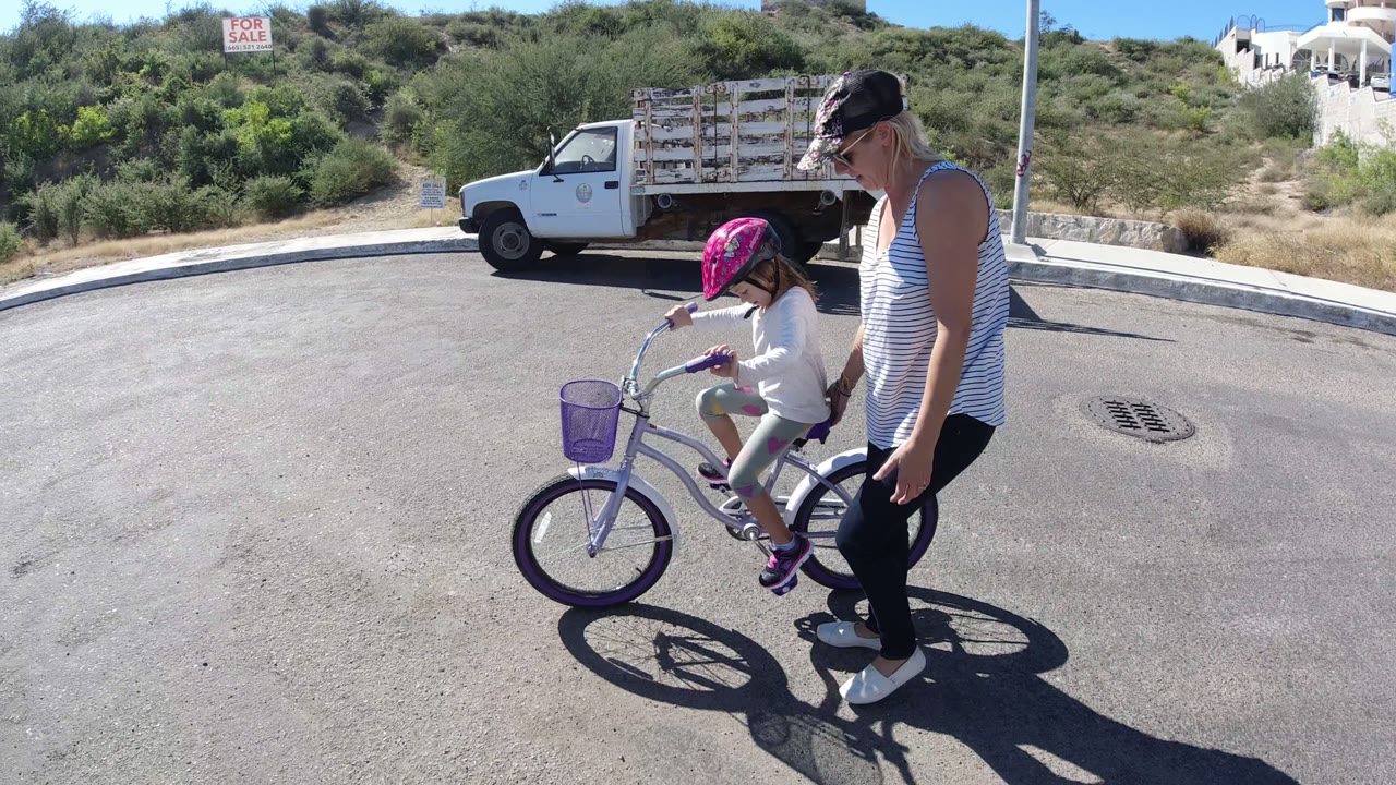 In San Jose del Cabo, the child pedals forward with both hands on the handlebars while the woman stays alongside her near the rear wheel.