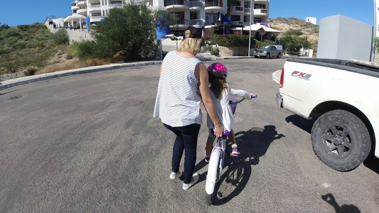 In San Jose del Cabo, the woman stays close to the child on the bicycle as they pass the rear side of a white pickup truck on a sunlit road with low hillside vegetation beyond.