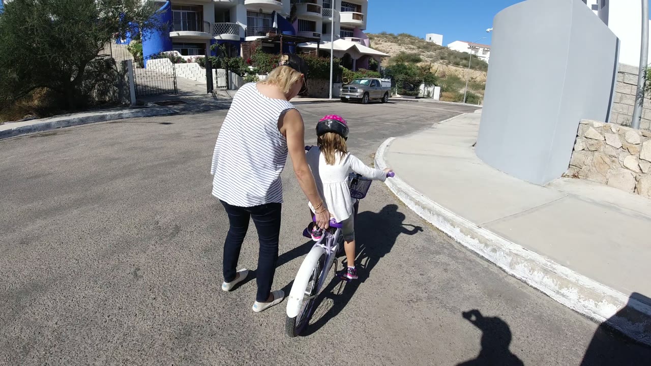 In San Jose del Cabo, Mexico, a woman walks beside a child on a purple bicycle near a curved curb, with white walls, parked cars, hillside plants, and residential buildings ahead.