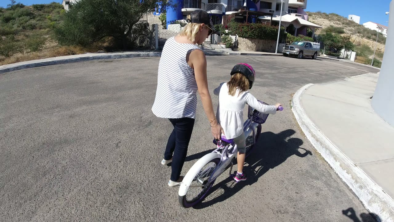The woman stays close to the child's rear wheel as the bicycle tracks toward the right-side sidewalk curve, with bright sun casting clear shadows on the asphalt.