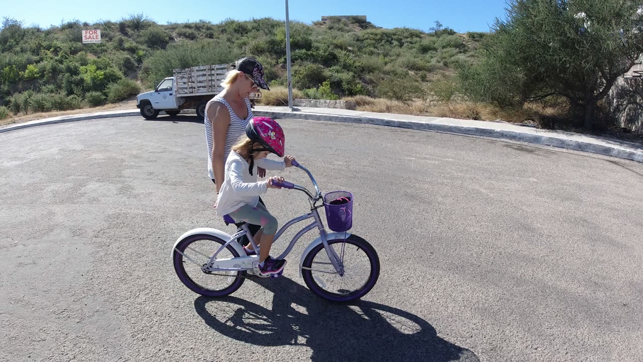 Woman walking beside a young girl on a small white bicycle.