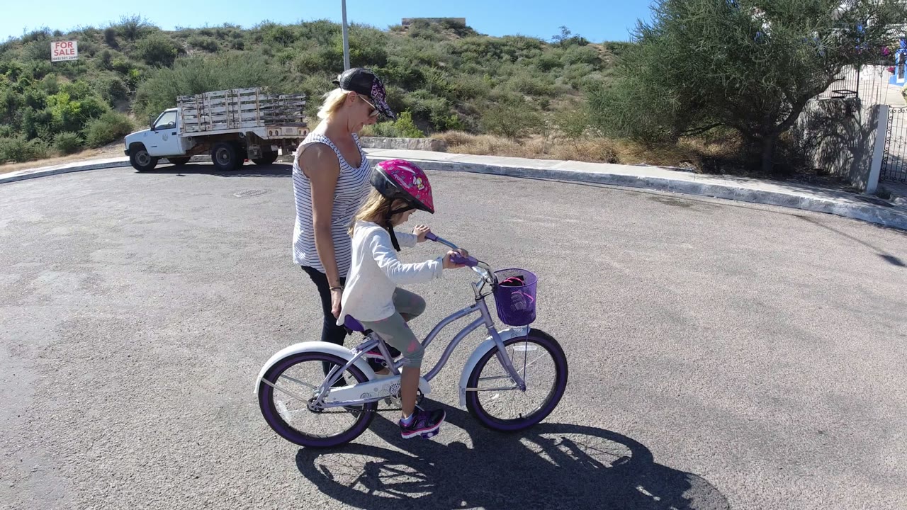 In San Jose del Cabo, Mexico, the girl pedals farther across the street with the woman beside her, a parked truck and green hillside visible behind them.