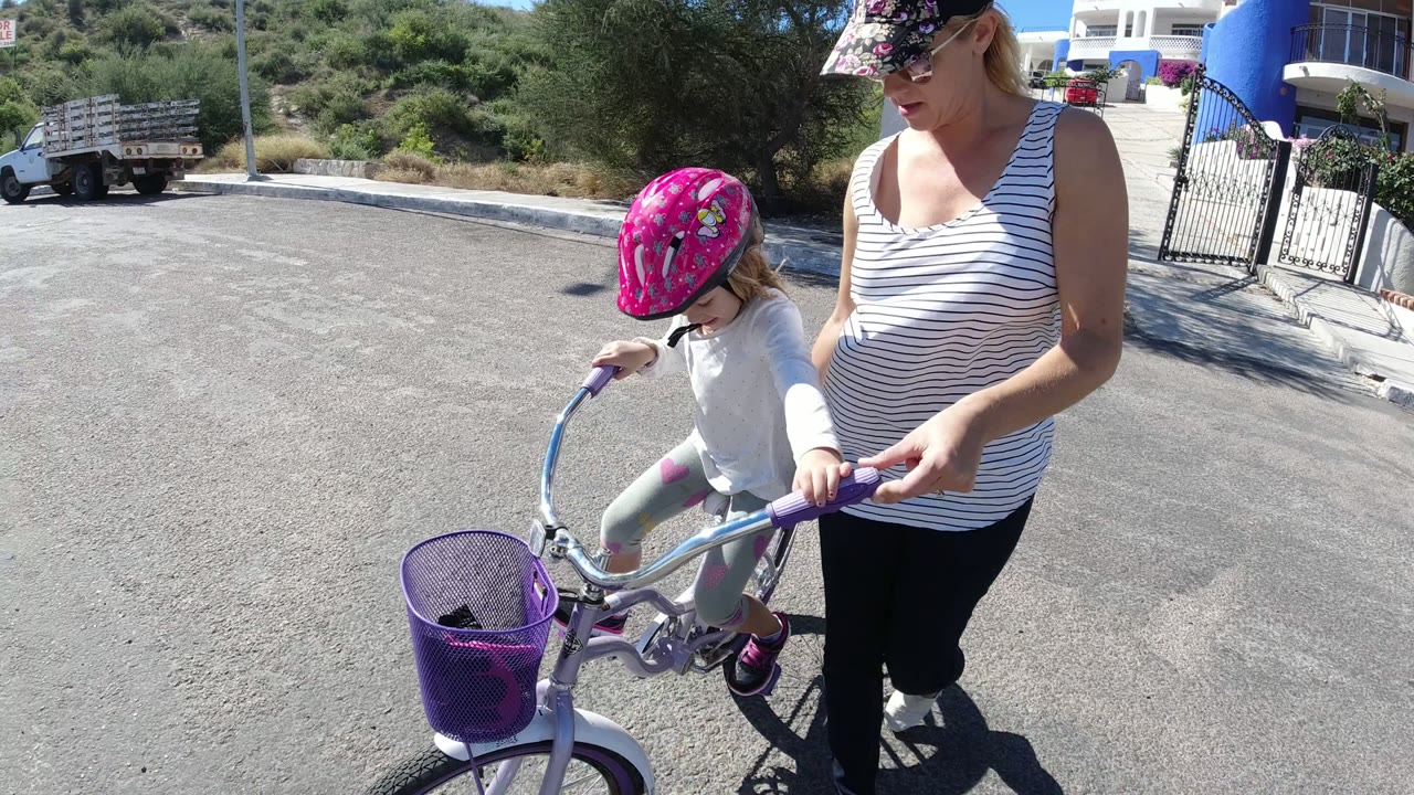 In San Jose Del Cabo, Mexico, a woman walks beside a small girl wearing a pink helmet on a purple bicycle near a paved roadside and low green hillside.