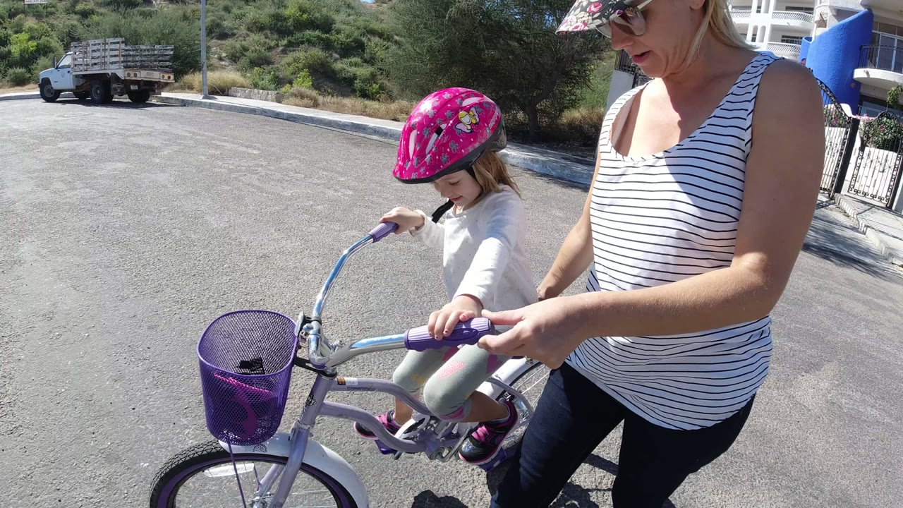 A closer side view in San Jose Del Cabo shows the child seated on the small purple bicycle while the woman keeps one hand near the handlebar on the sunlit pavement.