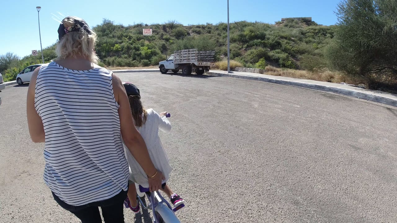 At 0:00 in San Jose del Cabo, Mexico, a woman walks behind and beside a child on a small bicycle, with parked vehicles and a green hillside ahead.