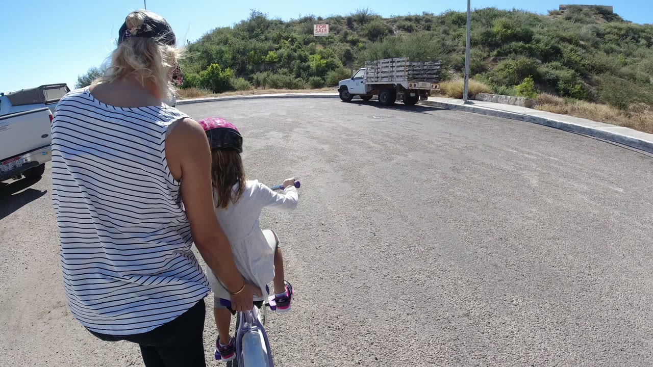 At 0:01, the woman and child continue through the paved lot, with a pickup truck close on the left edge and a flatbed truck near the hillside.