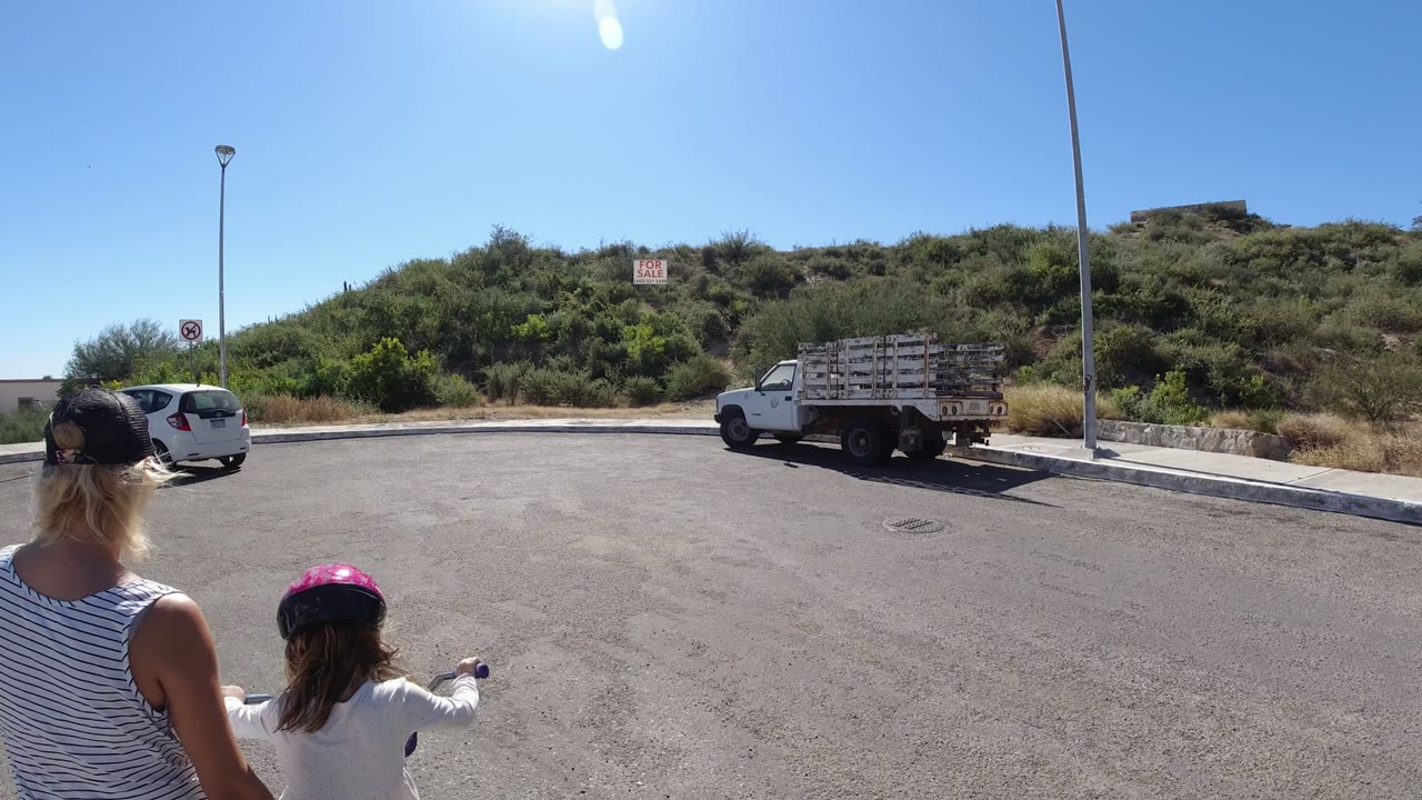 A woman and young child are partly cropped at the left edge in San Jose del Cabo, facing a sunny paved area with a hillside, streetlight, white van, and flatbed truck ahead.