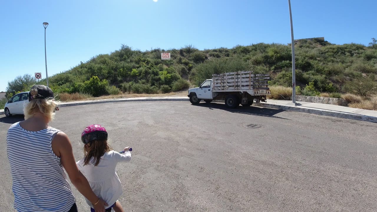 A mother and daughter are centered from behind in San Jose del Cabo, with the child in a pink helmet pointing across the pavement toward a white flatbed truck and brush-covered hill.