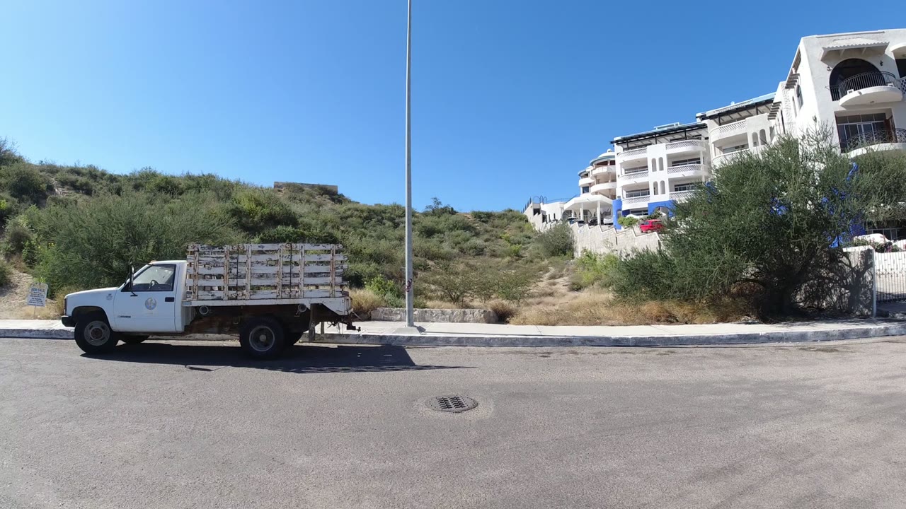 Wide street-level frame in San Jose del Cabo, Mexico showing a small white truck, hillside brush, white residential buildings, blue sky, and no visible people.